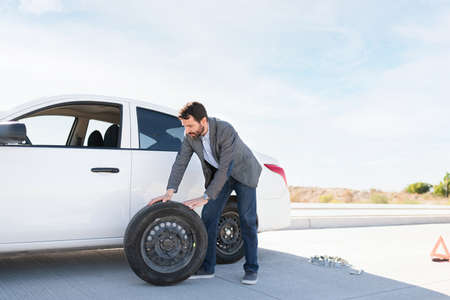 Latin man pulling the spare tire out of his car to change the flat one on the roadside