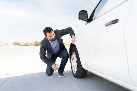 Hispanic man checking his vehicle for a flat tire. Man in his 30s with car problems parked on the side of the highwayの写真素材