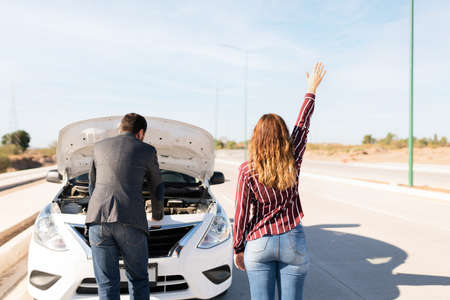 Couple with their car parked on the side of the road because of a flat battery. Young woman waving to get help to jump start their carの写真素材