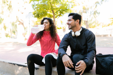 Handsome young man flirting and talking with a pretty woman that is drinking water. Fitness couple resting after working out in the parkの写真素材