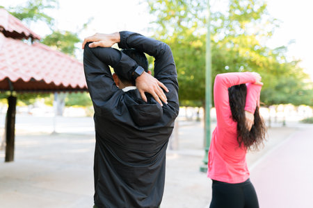 Fit man and woman in their 20s doing stretching exercises for the arms on the park. Rear view of a couple warming up before working outの写真素材
