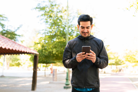 Hispanic handsome man in her 20s wearing sporty clothes is texting on his smartphone in the park