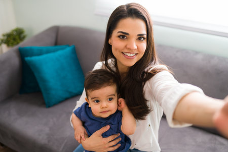 Pretty young woman with her baby boy taking selfie while sitting on sofa at home