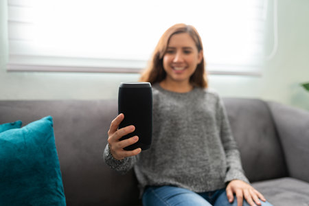 Happy caucasian young woman holding a new smart speaker and sitting on her couch at homeの写真素材