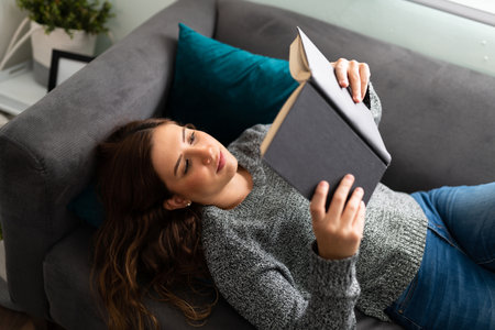 Attractive woman in her 20s seen from above, lying on the sofa living room and reading a bookの写真素材