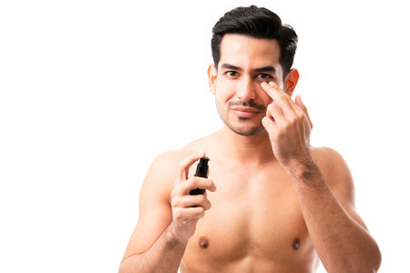 Hispanic man applying anti-aging cream under his eyes while making eye contact against white backgroundの写真素材