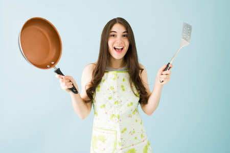 Caucasian woman in her 20s with an apron is happy and ready to start cooking. Young woman holding a frying pan and a spatulaの写真素材