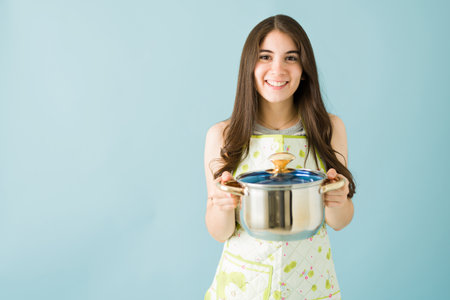 Portrait of a smiling young woman wearing an apron and holding a cooking pot with soup in front of a blue backgroundの写真素材
