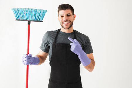 Portrait shot of an attractive young man with an apron and gloves. Hispanic guy is smiling and holding a broom because he finished all the houseworkの写真素材