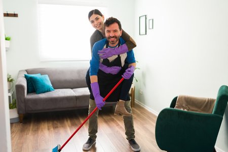 Cute young Latin couple cleaning their apartment together and woman piggyback riding her boyfriendの写真素材