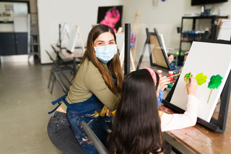Portrait of a hispanic female teacher with a face mask teaching a girl child how to paint with a...