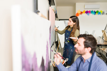 Side view of a beautiful young woman and attractive adult man taking a class in art school and painting with a brush on their canvasesの写真素材
