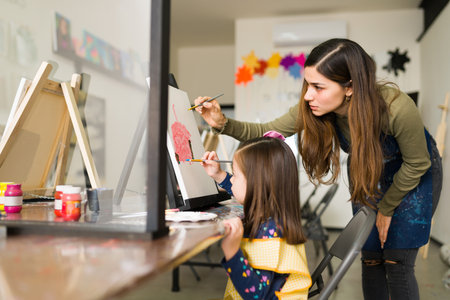 Attractive hispanic teacher is teaching a pretty child girl how to use the paint brush on a canvas during an art classの写真素材