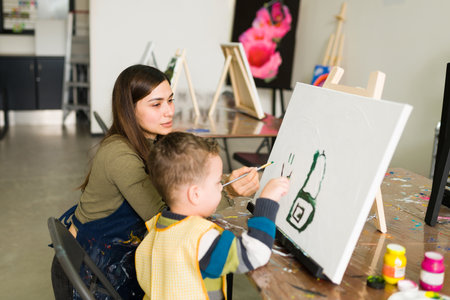 Hispanic female teacher is instructing a caucasian little boy to use a brush and do a painting on a blank canvas for an art classの写真素材