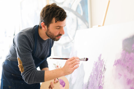 Attractive male artist in his 30s is giving the last touches of paint to the new colorful painting that he is doing on his art studioの写真素材