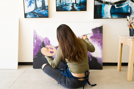 Latin female painter sitting on the floor and holding a big brush and a palette while giving the last touches of paint to her new artworkの写真素材