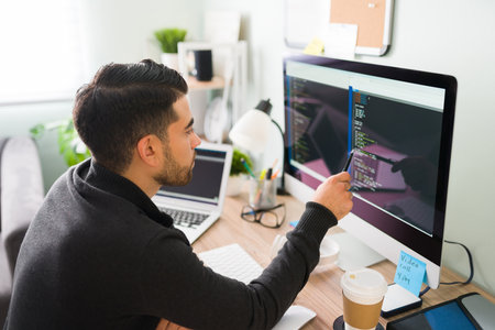 Rear view of a young software developer holding a pen pointing to the computer screen and is...