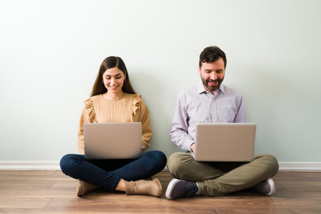 Attractive latin couple with laptops sitting together on the floor and looking for a partner on an online dating love websiteの写真素材