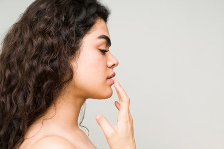Beautiful young hispanic woman touching her pink lips. Profile of a female beauty model with natural, clean skin and bare shouldersの写真素材