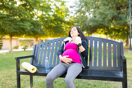 Tired pregnant woman looking exhausted while sitting on a bench park. Active woman resting after her workout exercisesの写真素材