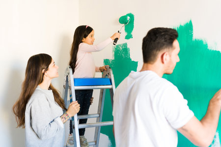 Happy latin family painting together the walls of their new home. Mom and dad applying green paint to the bedroom along with their smiling daughterの写真素材