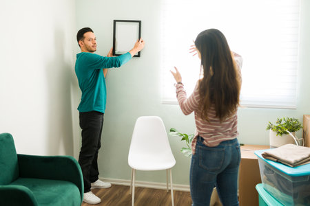 Girlfriend telling her boyfriend to move a picture frame in the living room wall. Couple decorating their stylish and modern apartmentの写真素材