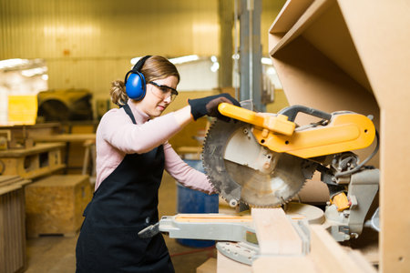 Caucasian young woman using hearing protection, glasses and gloves while using a bench saw. Female...
