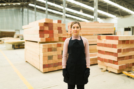 Pretty young woman looking at the camera while standing inside a wood warehouse. Portrait of a female worker in a woodshopの写真素材