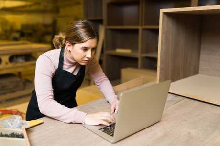 Pretty carpenter with an apron working on a laptop inside a big workshop. Female worker looking...