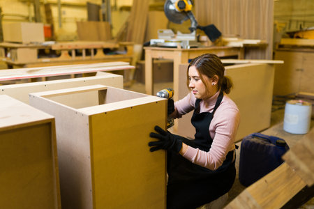 Female carpenter building a piece of wooden furniture in a workshop. Pretty worker drilling a screw with a power drill into a wood cabinetの写真素材