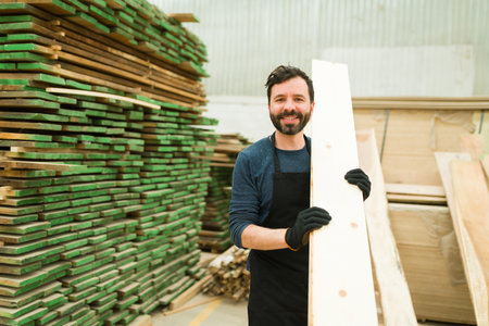 Portrait of a handsome latin worker choosing the best wooden boards. Carpenter standing next to a big stack of wood bars in a warehouseの写真素材