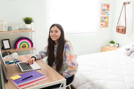 Happy teen girl typing in a laptop while sitting on the desk. Pretty caucasian teenager girl ready to do some homework in her bedroomの写真素材