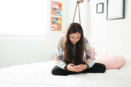 Good-looking teen girl sitting cross legged in bed and looking down at her smartphone. Happy teenager using social media and texting on her phoneの写真素材