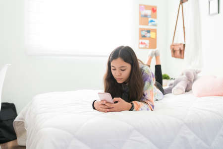 Caucasian teenage girl resting in her bedroom and texting on her phone. Adolescent girl lying on her stomach in bed and looking at her smartphoneの写真素材