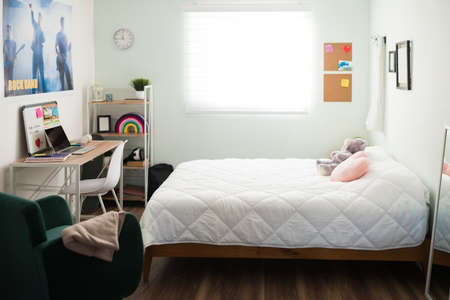 Beautiful teen bedroom with girly decoration. Bright adolescent's room with a sunny window, white bed, student desk, shelf, chair and a band poster on the wallの写真素材