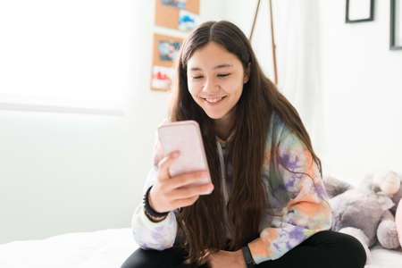 Smiling adolescent girl texting with her friends in a smartphone while sitting on her bedroom. Good-looking teenager girl chatting on a social media platformの写真素材