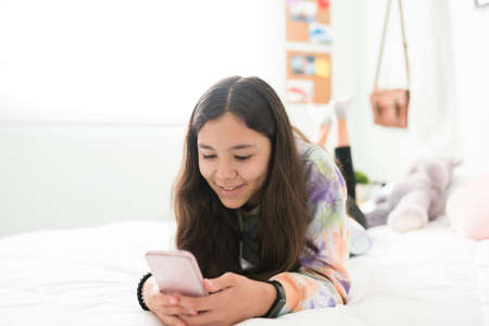 Smiling teen girl lying on her stomach in bed and texting on her phone. Adolescent girl relaxing in her bedroom and looking at social media photosの写真素材