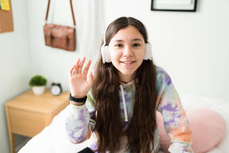 Beautiful caucasian teen girl waving hello during a video call with her friends and classmates. Teen girl talking online with friends during a video chatの写真素材
