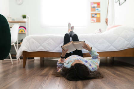 Studious adolescent girl reading a book and enjoying a story in her bedroom. Young teenage girl lying on the floor with her feet up on her bedの写真素材