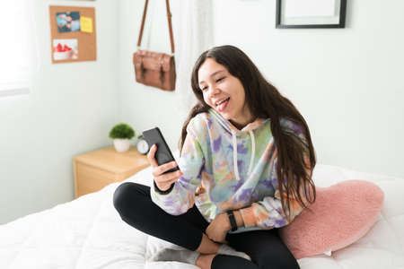 Funny teenager girl taking a selfie in her bedroom with her smartphone. Caucasian adolescent girl making a silly face with her tongue outの写真素材