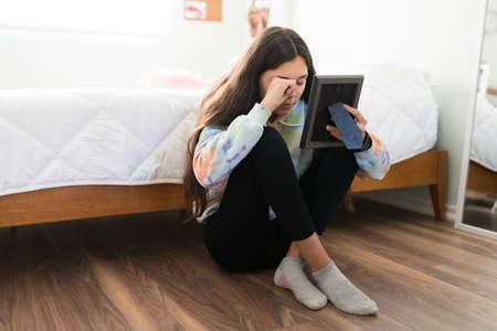 Sad caucasian teenage girl sitting on her bedroom floor. Heartbroken teen girl crying while looking at a photo frame of a loved oneの写真素材
