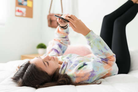 Good-looking adolescent girl lying in bed with her feet up in the wall. Caucasian teen girl scrolling through social media and texting on her smart phoneの写真素材