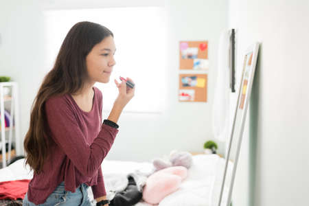 Profile of a girly teenager putting lipstick in front of the mirror. Beautiful teen girl putting makeup on inside her sunny and bright bedroomの写真素材