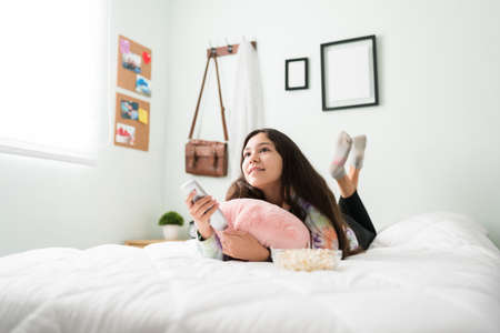 Happy teenager girl lying on her stomach in bed and holding the smart tv remote. Beautiful adolescent girl looking for a movie to watch in the bedroomの写真素材