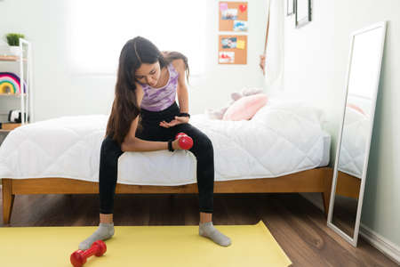 Active teenage girl sitting sitting on the bed and lifting weights. Beautiful adolescent girl exercising in her bedroom with dumbbellsの写真素材