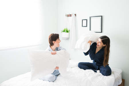 Good-looking young women with pajamas laughing and having a pillow fight in bed. Best friends...