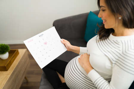 Side view of a caucasian pregnant woman holding and looking at a calendar. Expectant mother excited to mark a due date for the birth of her babyの写真素材