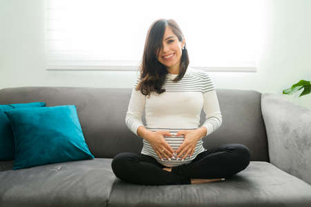 Attractive pregnant woman in her 30s with a beaming smile sitting on the couch and making a love heart sign with her hands in the bellyの写真素材