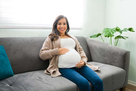Beautiful pregnant woman in her 30s wearing casual clothes and sitting on her home living room. Happy expectant mother resting on the couchの写真素材