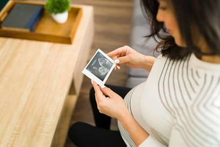 High angle of a loving pregnant woman smiling and looking happily at the print echography of her new baby while sitting in her home living roomの写真素材